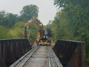 Un travailleur portant un casque de sécurité et des lunettes de protection surveille un marteau hydraulique Grizzly MG-60 monté sur une excavatrice CAT 329F flambant neuve sur chenilles en train de réparer un pont ferroviaire.