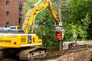 A yellow Komatsu excavator is parked at a road construction site, with a warning sign and trees in the background. The logo and phone number of the company "Inter Chantiers" are visible on the excavator.