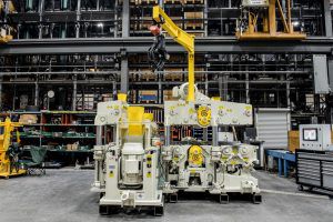 Une raboteuse industrielle Gilbert Mass Timber Planer est installée dans un atelier spacieux. Elle est de couleur crème et grise, avec des accents jaunes sur le bras de levage et les panneaux de commande. La raboteuse est entourée de divers outils et équipements et un chariot élévateur jaune est visible en arrière-plan.