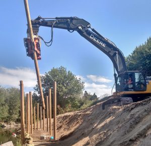 Une pelle mécanique John Deere grise équipée d'un vibrofonceur Gilbert MG-90 enfonce un pieu en bois dans le sol, le long d'une berge, pour créer une structure de soutènement.