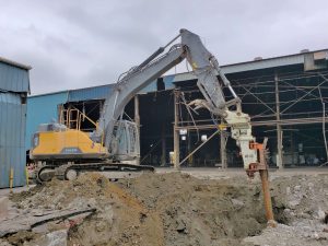 A powerful Grizzly MG-90 combined with a Volvo EC380L excavator rips out wooden piles within seconds, with a building under demolition or renovation in the background.