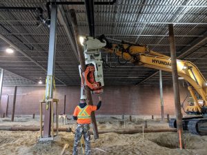 Un vibrofonceur MG-90 Gilbert, fixé à une excavatrice Hyundai, vibre et s'enfonce dans le sol pour préparer une fondation à l'intérieur d'un bâtiment. Un homme en casque et veste réfléchissante orange guide l'opérateur de l'excavatrice avec des gestes précis. Des rubans jaunes 