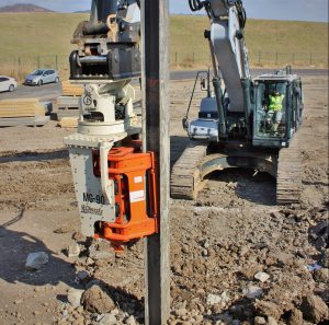 Une pelle hydraulique équipée d'un vibrateur de pieux latéral Gilbert Grizzly MG-90 enfonce un pieu métallique en H dans le sol, sur un chantier de construction.