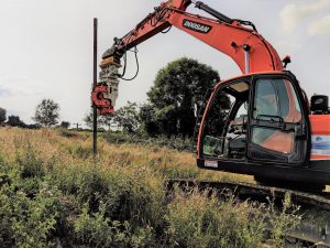 Une pelle mécanique Doosan rouge vif est utilisée pour enfoncer un pieu en acier dans le sol avec un Vibrofonceur à prise latérale Gilbert Grizzly MultiGrip MG-60. Le conducteur de la pelle, visible dans la cabine, manœuvre soigneusement le pieu en place à l'aide d'un marteau-pilon. L'équipement est situé dans un champ herbeux, peut-être pour la construction d'une nouvelle structure.