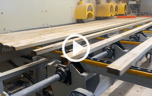 An industrial planer with multiple cutter heads is planing wooden boards. The boards are transported on a roller conveyor and pass under the rotating cutter heads. The machine is equipped with yellow safety guards.