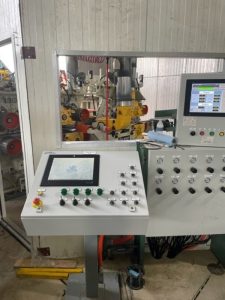 Un panneau de contrôle industriel avec deux écrans et divers boutons et voyants. Le panneau de contrôle est de couleur grise et blanche, et il est monté sur un support métallique. Derrière le panneau de contrôle, on peut voir une machine industrielle avec des rouleaux et des tuyaux.